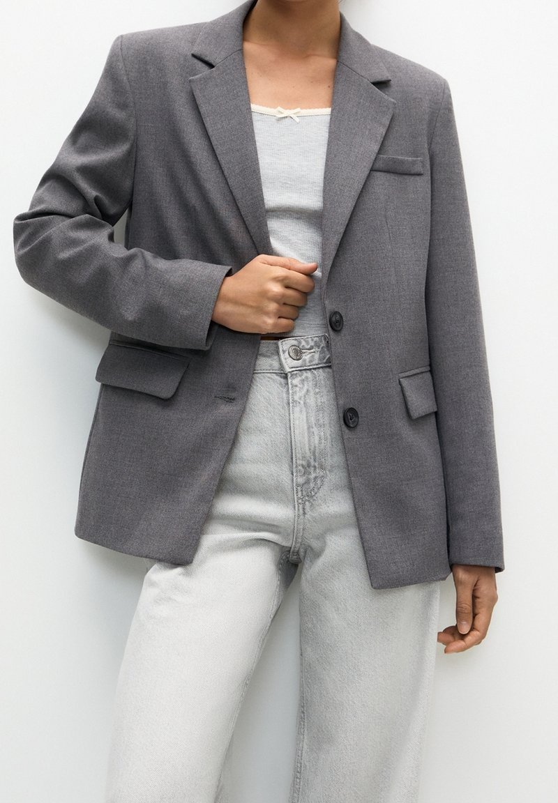 Personne portant un jean gris clair, un débardeur blanc et un blazer gris, ajustant le blazer d'une main.