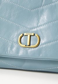 Šviesiai mėlyna quilted odinė rankinė su auksiniu logotipu užsegimu ir balta siuvinėjimu, pabrėžianti lygų paviršių ir struktūrizuotą dizainą.