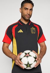 Camisa polo preta com detalhes em vermelho e amarelo, apresentando um colarinho com fecho zip e logo. O modelo segura uma bola de futebol branca com detalhes em preto e verde.