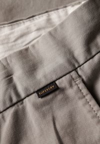 Grå stof med en struktureret tekstur, med et sort mærke der står "SUPERDRY" i gyldne sting. Pænt foldet design.