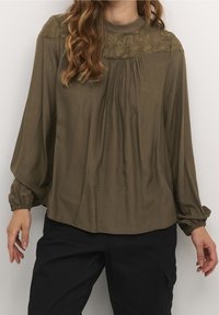 Blouse vert olive avec un col haut, des détails en dentelle florale sur les épaules et un devant plissé. Manches longues et légères avec des poignets froncés.
