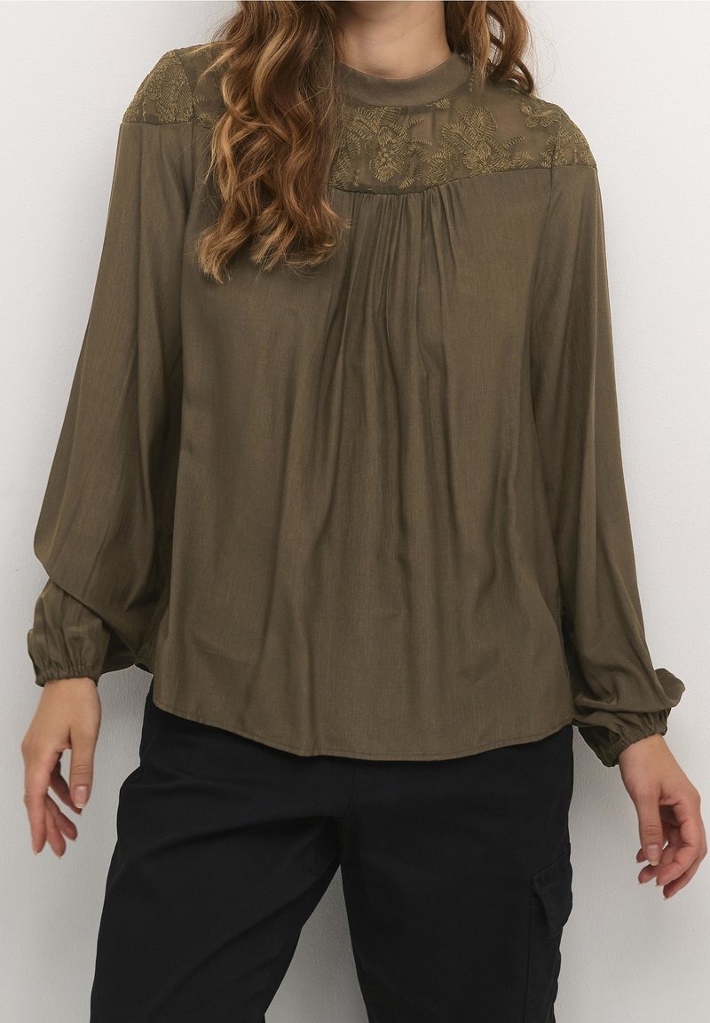 Blouse vert olive avec un col haut, des détails en dentelle florale sur les épaules et un devant plissé. Manches longues et légères avec des poignets froncés.