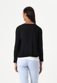 Zwart lichtgewicht sweater met driekwartmouwen, textuur gebreid, cropped ontwerp, zijsplits, gedragen over een wit topje en lichtblauwe broek.