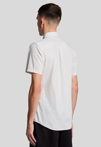 Ricerca a maniche corte, camicia con colletto color beige chiaro con un motivo a quadri. Presenta uno yoke posteriore dritto e spacchi laterali per facilitare il movimento.