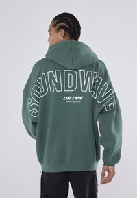 Πράσινη κουκούλα φούτερ με την ένδειξη "SOUNDWAVE" εκτυπωμένη στην πλάτη με μεγάλους λευκούς χαρακτήρες; διαθέτει χαλαρή εφαρμογή και ριμπ μανσέτες.