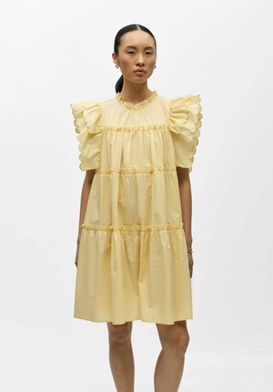 Giovane donna che indossa un abito a balze giallo pallido con maniche corte arricciate a scallop e collo alto, in piedi contro uno sfondo bianco uniforme.