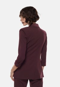 Donna con capelli corti e mossi, che indossa un blazer aderente bordeaux e pantaloni abbinati, rivolta all'indietro contro uno sfondo chiaro e semplice.