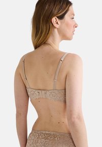 Soutien-gorge en dentelle beige avec motif floral, bretelles réglables et fermeture à crochets au dos. Texture lisse avec une finition délicate.