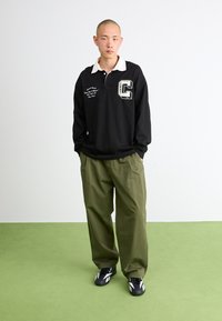 Polo a maniche lunghe nera con colletto bianco, dettagli ricamati e grande logo "C"; abbinato a pantaloni larghi verde oliva e sneakers nere.