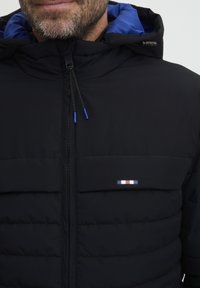 Črna pufasta jakna z modrim notranjim oblogo, ki ima zadrgo, nastavljive vrvice in majhen logotip v rdeči, beli in modri barvi.