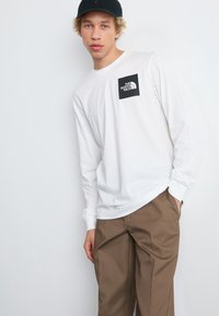 Long-sleeve white shirt with a black logo patch from The North Face, paired with khaki pants. Features a relaxed fit and crew neckline.