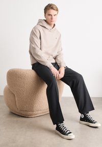 Beige kapucnis pulóver elülső zsebbel, fekete nadrág és fekete magasszárú sneakers. Ül egy texturált, világosbarna, kerek széken.