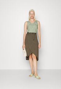 Top de alças verde claro com bordados florais na gola, combinado com uma saia envolvente castanha com um laço na cintura. Acessórios incluem uma bolsa bege e sandálias verdes.