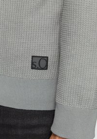 Lichtgrijze sweater met een textuurpatroon, met een ribgebreide zoom en manchetten. Een klein zwart label met "s.O" is zichtbaar aan de zijkant.