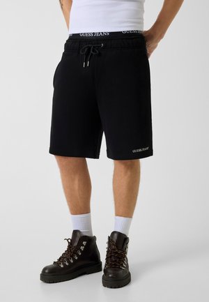 Mannequin masculin portant un short noir Guess Jeans à cordon, des chaussettes blanches, des bottes de randonnée marron foncé et un haut blanc sans manches, debout les mains derrière le dos.