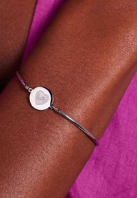 Zilveren manchetarmband met een ronde bedel met een hartontwerp. Gladde metaaltextuur, gedragen op een bruine huidtoon. Achtergrond van roze stof.