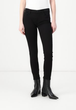 Mujer con pantalones negros ajustados y botines negros de tacón, de pie frente a un fondo liso y claro.