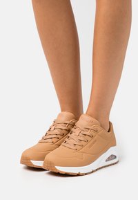 Beige Sportschuhe mit strukturiertem Obermaterial, runder Zehenpartie, Schnürsenkeln und weißer Zwischensohle; verfügen über eine sichtbare Luftpolsterung.