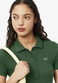 Grünes, geripptes Polo-Shirt mit kurzen Ärmeln, mit Kragen und kleinem Lacoste-Krokodil-Logo auf der Brust. Glatte Textur.