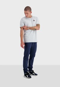 Grijze katoenen t-shirt met een logo op de borst, gecombineerd met marineblauwe sweatpants en zwarte sportschoenen met witte accenten.