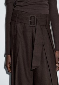 Gros plan sur un pantalon taille haute de couleur brun foncé, sur mesure, avec une ceinture en tissu et une boucle rectangulaire, porté avec un haut à manches longues assorti.