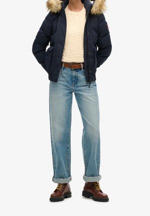 Personne portant une doudoune bleu marine avec capuche en fourrure, un pull en maille crème, un jean bleu clair retroussé, une ceinture marron et des bottes marron lacées.