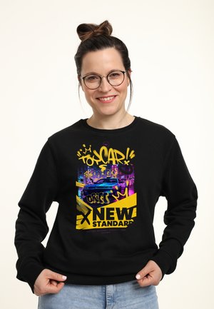 Kvinde med briller og knold frisurer smilende, iført sort sweatshirt med farverig graffiti-stil bil og teksten "New Standard" design.