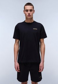 Black cotton t-shirt with short sleeves, featuring a small horizontal logo patch in the center. Styled with dark shorts.
