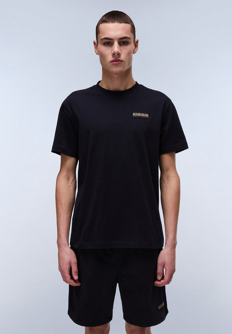 Black cotton t-shirt with short sleeves, featuring a small horizontal logo patch in the center. Styled with dark shorts.
