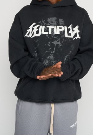 Person wearing a black hoodie with a stylized "MULTIPLY" logo and graphic face, hands clasped in front, paired with gray sweatpants.