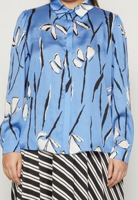 Donna che indossa una blusa blu con motivi floreali astratti in bianco e nero e una gonna a righe bianche e nere.