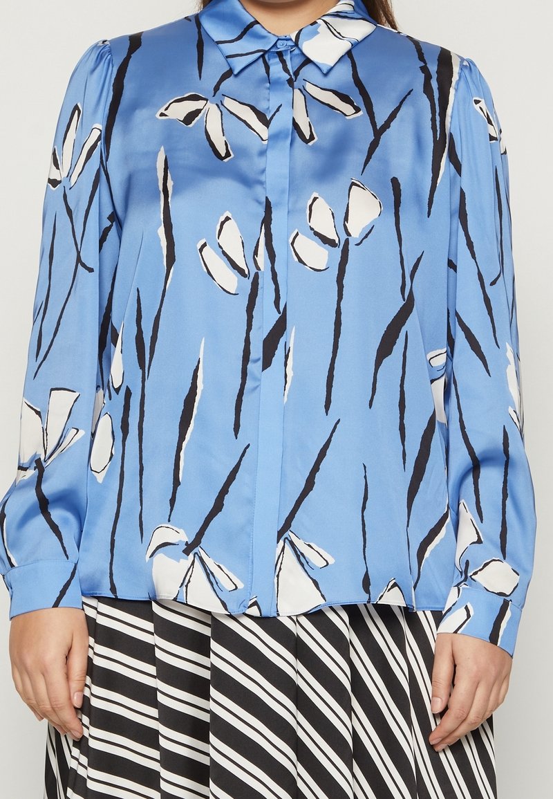 Donna che indossa una blusa blu con motivi floreali astratti in bianco e nero e una gonna a righe bianche e nere.