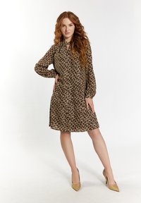 Braunes Seidenkleid mit einem kreisförmigen Muster, Schleifenkragen und langen Ärmeln. Trägt beiges spitz zulaufendes Paar Schuhe. Das Model posiert mit einer Hand auf der Hüfte.