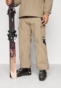 Μπεζ απομονωμένα παντελόνια χιονιού με υφή, πλαϊνές τσέπες cargo και μαύρες λεπτομέρειες, συνδυασμένα με μαύρες μπότες χιονιού και ένα μωβ σκι.