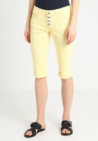 Pantalones amarillos cortos con cierre frontal de cinco botones, puños doblados y una textura suave, combinados con sandalias negras de tiras.
