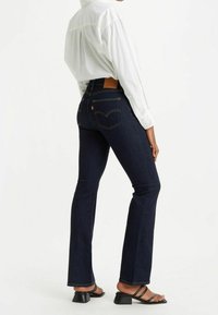 Jean en denim bleu foncé à taille haute, jambes évasées, avec des coutures jaunes et une étiquette en cuir marron au dos portant un logo. Associé à une chemise blanche.