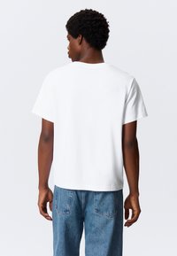 Camiseta branca de algodão com mangas curtas, decote redondo e corte descontraído. Usada com jeans denim azul claro.