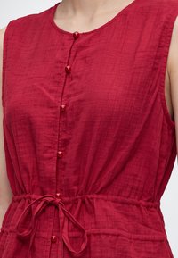 Robe rouge sans manches avec encolure ronde, boutons recouverts de tissu sur le devant et ceinture nouée à la taille.