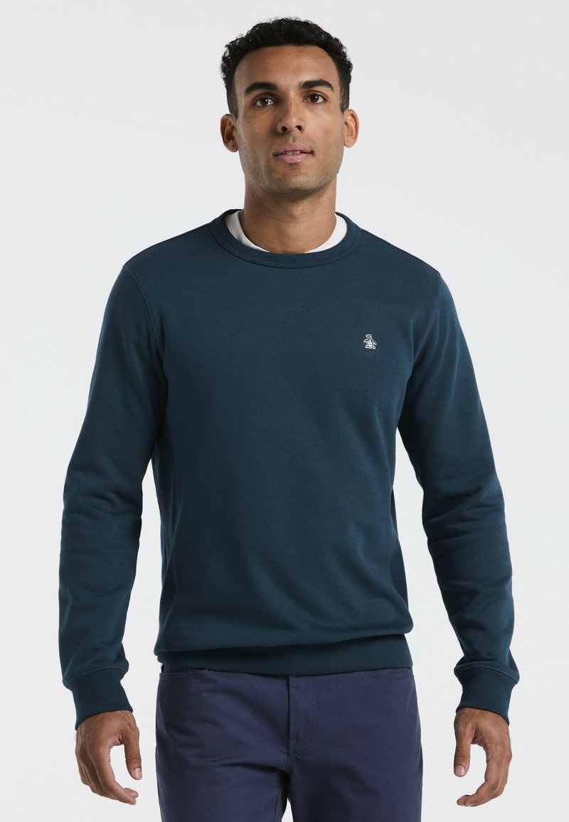 Man staat rechtop en kijkt naar voren, gekleed in een donkerblauw sweatshirt met ronde hals over een wit shirt en marineblauwe broek, tegen een witte achtergrond.