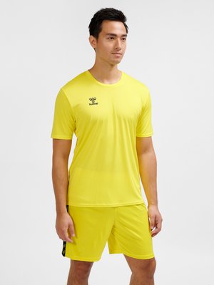 Mand stående iført gul kortærmet sportstrøje og matchende shorts med lille sort brandlogo på brystet, neutral baggrund.