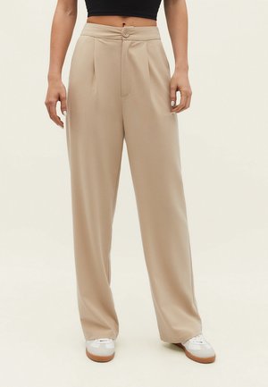 Pantalons beige à jambes larges avec une texture lisse, dotés d'une fermeture à bouton, d'un pli central et d'une taille haute. Associés à des baskets blanches.
