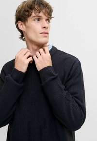 Suéter de punto azul marino con una superficie texturizada y puños acanalados. Diseño de cuello redondo, llevado sobre una camisa con cuello de rayas azules y blancas.