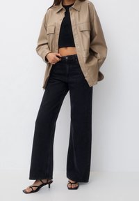 Veste-chemise en similicuir beige avec deux poches avant, associée à un jean noir taille haute à jambes larges et des sandales à talons noires avec lanières.