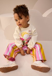 Sweat-shirt blanc avec broderie colorée d'animaux et de soleil ; pantalon large rayé en rose, jaune et orange ; chaussures blanches avec semelles texturées.