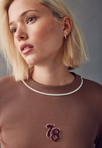 Camisa marrón de punto acanalado con acento de cuello blanco, con detalle bordado "78" en burdeos. Primer plano del escote y el rostro del modelo.