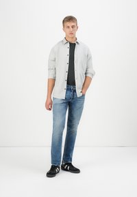 Giovane in piedi, con una camicia grigia chiaro a bottoni sopra una t-shirt nera, jeans blu e scarpe da ginnastica nere con strisce bianche, su sfondo bianco.