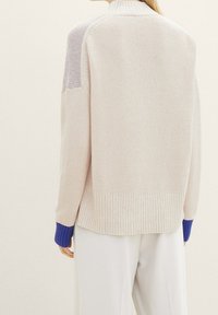 Person in einem beigen, strukturierten Strickpullover mit blauen Rippenbündchen und weißen, locker sitzenden Hosen, von hinten gezeigt.