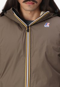 Giacca impermeabile marrone con zip e strisce multicolori, fodera interna morbida e logo sul petto.
