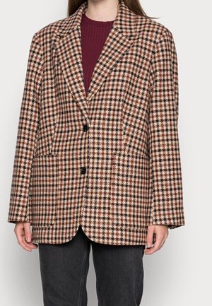 Woman wearing a beige, brown, and red checkered blazer over a burgundy ribbed top and black pants, standing against a plain light background.