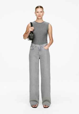 Graues ärmelloses Top kombiniert mit hochgeschnittenen, lockeren grauen Jeans. Das Model hält eine kleine schwarze Handtasche und trägt schwarze High Heels.